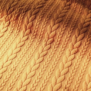Peut inclure: Gros plan sur un tissu textur&eacute; jaune dor&eacute;. Le motif pr&eacute;sente des rang&eacute;es de motifs r&eacute;p&eacute;t&eacute;s de feuilles ou d'&eacute;pis de bl&eacute; stylis&eacute;s. Le tissu a un aspect doux et matelass&eacute;, avec le motif en relief cr&eacute;ant des ombres et de la profondeur.