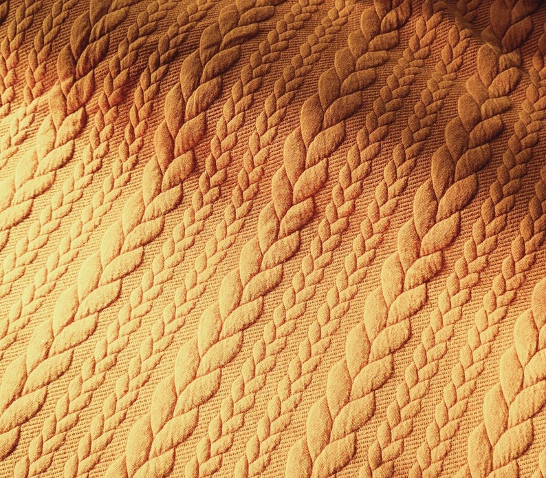 Peut inclure: Gros plan sur un tissu textur&eacute; jaune dor&eacute;. Le motif pr&eacute;sente des rang&eacute;es de motifs r&eacute;p&eacute;t&eacute;s de feuilles ou d'&eacute;pis de bl&eacute; stylis&eacute;s. Le tissu a un aspect doux et matelass&eacute;, avec le motif en relief cr&eacute;ant des ombres et de la profondeur.