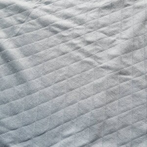 Peut inclure: Gros plan sur un tissu gris avec un motif losange matelass&eacute;. Le tissu a un aspect doux et textur&eacute; avec des lignes diagonales et des ombres subtiles, cr&eacute;ant une sensation de profondeur. Le ton g&eacute;n&eacute;ral est neutre et apaisant.