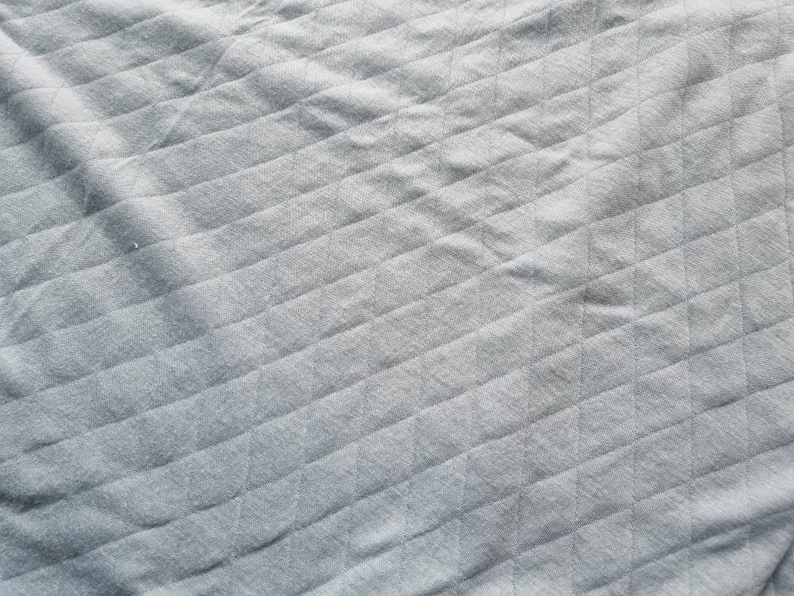 Peut inclure: Gros plan sur un tissu gris avec un motif losange matelass&eacute;. Le tissu a un aspect doux et textur&eacute; avec des lignes diagonales et des ombres subtiles, cr&eacute;ant une sensation de profondeur. Le ton g&eacute;n&eacute;ral est neutre et apaisant.