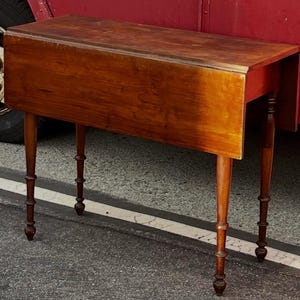 May include: A vintage wooden drop-leaf table with a warm, reddish-brown finish. The table features a rectangular top and four slender, turned legs. The drop-leaf is extended, and the table is set on an asphalt surface.