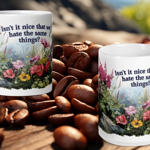 May include: Two white ceramic mugs with a floral design and the text "Isn't it nice that we hate the same things?" printed on them. The mugs are sitting on a bed of coffee beans.