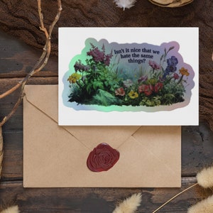 May include: A sticker with a holographic finish featuring a colorful floral illustration and the text "Isn't it nice that we hate the same things?"