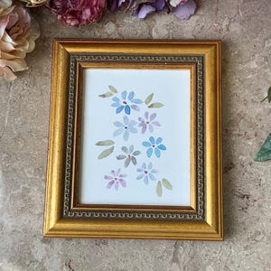 May include: A framed watercolour painting of blue and purple flowers on a white background. The frame is gold with a decorative border.