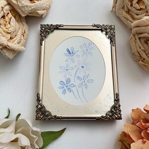 May include: A framed watercolour painting of blue flowers on a white background. The frame is silver with ornate details.