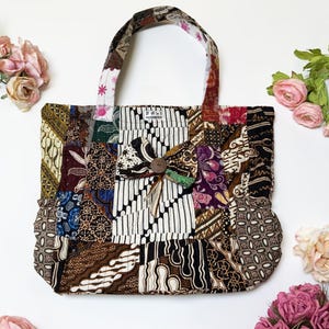 Peut inclure: Un sac fourre-tout en patchwork avec un motif batik brun, noir et blanc. Le sac a un nœud en tissu identique et un bouton en bois au centre. Le sac porte une étiquette indiquant "2111 INDONÉSIE".