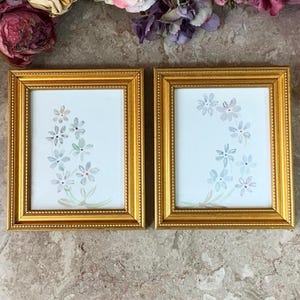 May include: Two gold-framed pictures with white mounts and watercolour paintings of blue flowers. The frames have a beaded edge.