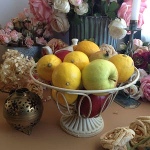 May include: A white metal fruit bowl filled with lemons, apples, and a red apple. The bowl is on a brown surface with a gold decorative object and pink flowers in the background.