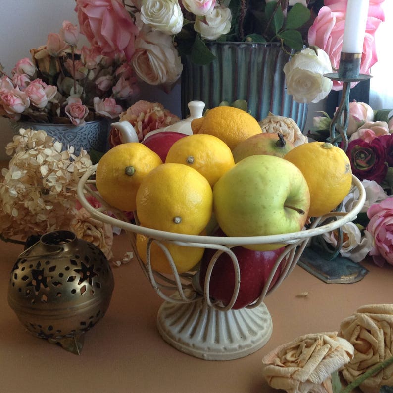 May include: A white metal fruit bowl filled with lemons, apples, and a red apple. The bowl is on a brown surface with a gold decorative object and pink flowers in the background.