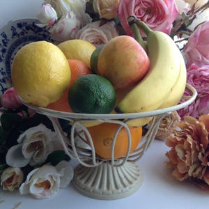 May include: A white wire fruit bowl with a pedestal base holds a variety of fruit, including lemons, limes, oranges, apples, and bananas. The bowl is surrounded by pink and white roses.