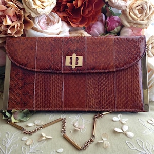 Peut inclure: Une pochette en cuir de serpent brun avec une cha&icirc;ne dor&eacute;e et un fermoir dor&eacute;. La pochette est ferm&eacute;e et pos&eacute;e sur une surface beige avec des fleurs blanches.