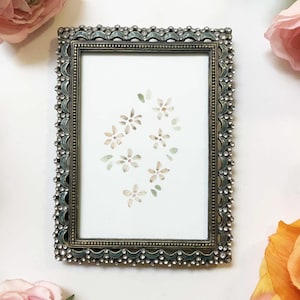 May include: An antique-style picture frame with a floral watercolour painting. The frame is dark silver with a beaded edge and small, clear crystals. The painting shows small, light brown and green flowers on a white background.