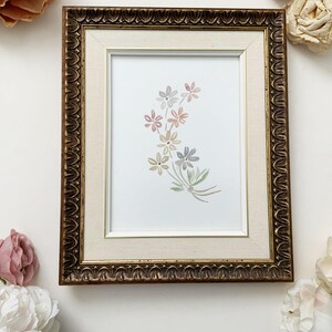 May include: A framed watercolour painting of a bunch of pastel-coloured flowers. The flowers are painted in shades of pink, yellow, and blue, and they are arranged in a loose, flowing style. The frame is a simple gold frame with a white mount.