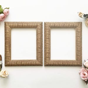 May include: Two square, ornate picture frames with a gold finish and a leaf pattern. The frames are empty, with a white background visible. Pink and cream-coloured flowers are arranged around the frames.