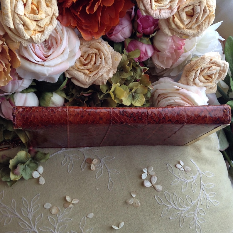 Peut inclure: Un journal reli&eacute; en cuir marron avec une couverture textur&eacute;e, semblable &agrave; du cuir de serpent. Le journal repose sur une surface en tissu blanc avec un motif floral.