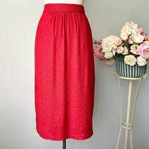 May include: A bright red skirt made from textured material. The skirt features a high waist and a straight cut. A floral display in a decorative pot is on a white metal stand in the background.