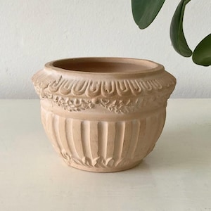 May include: A beige terracotta planter with an embossed design. The pot has vertical grooves and a decorative band of floral and scrollwork around the rim. Suitable for indoor or outdoor use, the pot is a classic design.