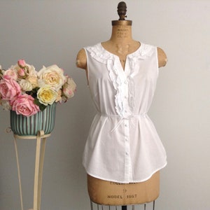 May include: A white sleeveless blouse with a ruffled neckline and a button-up front. The blouse has a cinched waist with a bow tie detail. The blouse is on a dress form.