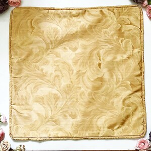 May include: Square cushion in a gold hue, featuring an elaborate, swirling leaf design. The fabric has a slight sheen, and the edges are trimmed with a darker gold border. The pattern is symmetrical. This cushion is likely for decorative purposes.