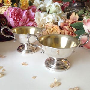 May include: Two silver serving bowls with ornate handles and a pedestal base. The bowls are polished and reflect light.