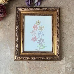 May include: A gold-coloured picture frame with a white mount and a watercolour painting of a bouquet of pink, purple, and blue flowers.
