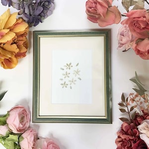 May include: A framed floral watercolour painting with a green and gold frame. The artwork features delicate, light-coloured flowers on a white background. The frame is surrounded by various colourful artificial flowers, including roses and tulips.