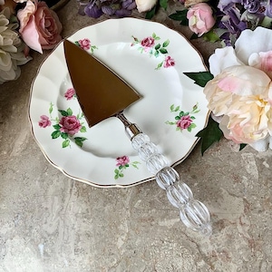May include: A gold cake server with a clear, beaded handle sits on a white plate. The plate is decorated with a floral pattern of pink roses and green leaves. The plate has a gold trim.