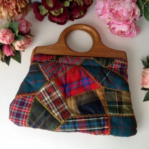 May include: A patchwork handbag with a wooden handle. The bag is made of different plaid fabrics in shades of red, green, blue, and yellow. The handle is a single, large loop.