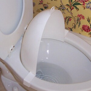 以下が含まれることがあります： 白い便器で蓋が開いており、白い飛沫ガードが付いています。 便器は花柄の壁紙を背景に設置されています。 便座も白です。