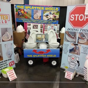 以下が含まれることがあります： 「The Splatter Shield」トイレ製品の展示ブース。ブースには、製品情報、画像、および「STOP DOING THIS」というテキストが記載された看板があります。3つのトイレを載せたカートが中央にあります。