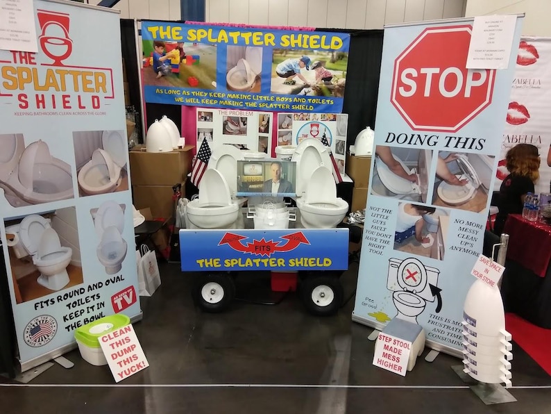 以下が含まれることがあります： 「The Splatter Shield」トイレ製品の展示ブース。ブースには、製品情報、画像、および「STOP DOING THIS」というテキストが記載された看板があります。3つのトイレを載せたカートが中央にあります。