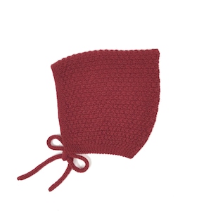May include: A deep burgundy knitted bonnet featuring a textured stitch pattern throughout the main body. The headwear includes a smooth, solid-coloured border along the front edge and two matching knitted ties attached at the base for fastening.