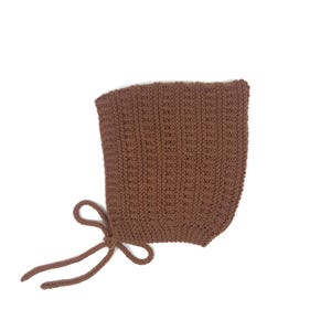 May include: A brown knitted bonnet with a textured pattern. The bonnet has a rounded shape and features long ties for securing under the chin. The colour is a warm, earthy brown, suitable for babies and young children.