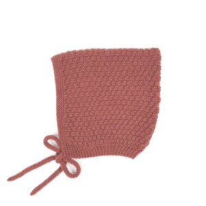 Può includere: Un cappello lavorato a maglia di colore rosa antico, con un motivo a punto strutturato. Il cappello ha una forma arrotondata e lacci per l'allacciatura. Il colore tenue e il design semplice suggeriscono un articolo per neonati o bambini. Il cappello è alto circa 20 cm.