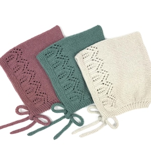 Può includere: Tre cappellini per neonati lavorati a maglia in rosa antico, verde acqua e crema. Ogni cappellino presenta un bordo in pizzo a motivo di foglie sul bordo anteriore e si lega con un nastro abbinato. I cappellini sono esposti su sfondo bianco.