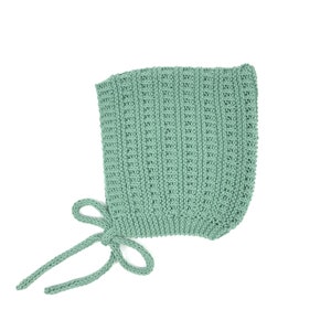 Può includere: Un cappellino da neonato lavorato a maglia verde salvia con un motivo a nido d'ape strutturato. Il cappellino ha una forma arrotondata e si lega con un fiocco sotto il mento. Il colore tenue e la lavorazione delicata suggeriscono un articolo fatto a mano.