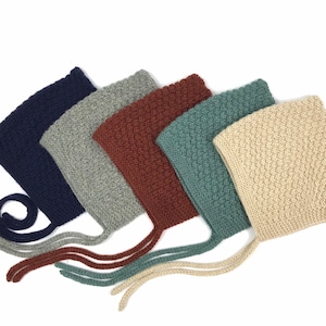 Può includere: Una collezione di sei cappellini per neonati lavorati a maglia in vari colori, tra cui blu navy, grigio, bordeaux, verde acqua e crema. Ogni cappellino presenta un motivo a maglia testurizzato e lacci per l'allacciatura. I cappellini sono disposti su sfondo bianco.