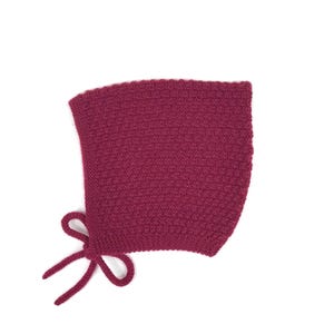 Op de afbeelding: Een diep magenta mutsje gemaakt met een gestructureerd breipatroon. Het hoofddeksel heeft een ronde vorm die het gezicht omlijst en is voorzien van twee bijpassende gebreide koordjes aan de onderste hoek, die samen zijn geknoopt.