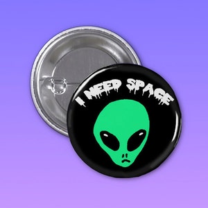 May include: A black and white button with a green alien face and the text "I NEED SPACE" in white dripping letters.