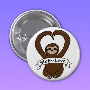 May include: A white button featuring a brown sloth forming a heart shape with its arms. The sloth has a smile and the text "Sloth Love" is displayed on a banner beneath it.