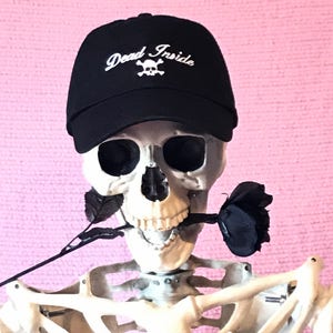 May include: A black baseball cap with a white skull and crossbones and the text "Dead Inside" embroidered on the front. The cap is worn by a skeleton holding a black rose.