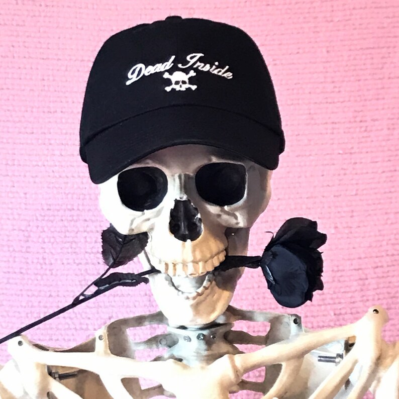 May include: A black baseball cap with a white skull and crossbones and the text "Dead Inside" embroidered on the front. The cap is worn by a skeleton holding a black rose.