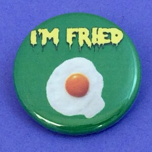 May include: A green button with the text "I'M FRIED" in yellow dripping letters. A white fried egg with a yellow yolk is in the center of the button.