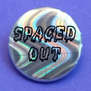 Puede incluir: Un botón holográfico plateado con el texto "SPACED OUT" en letras negras que gotean.