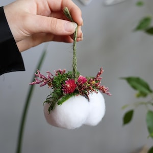 Könnte beinhalten: Handgefertigter weißer Filzschmuck, verziert mit einem Cluster aus leuchtend rosa und roten Blumen, grünem Moos und zarten Zweigen. Der Schmuck wird von einer natürlichen Schnur aus Jute gehalten, was eine charmante, rustikale Ästhetik erzeugt. Einzigartige Blumendekoration.