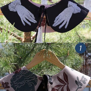 May include: Two decorative detachable collars on a wooden hanger. One is black with white hand illustrations, the other is tan with a dark green bust and floral design. Both have black ties.