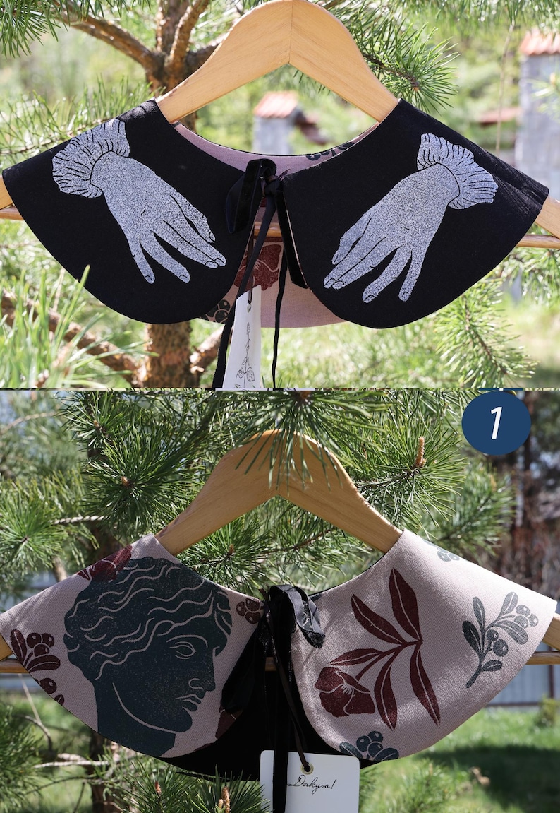 May include: Two decorative detachable collars on a wooden hanger. One is black with white hand illustrations, the other is tan with a dark green bust and floral design. Both have black ties.