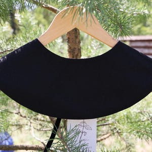 May include: A black, rounded collar, displayed on a wooden hanger. The collar has a smooth texture and a curved shape. The background features green foliage and a glimpse of a building. The collar is a fashion accessory.