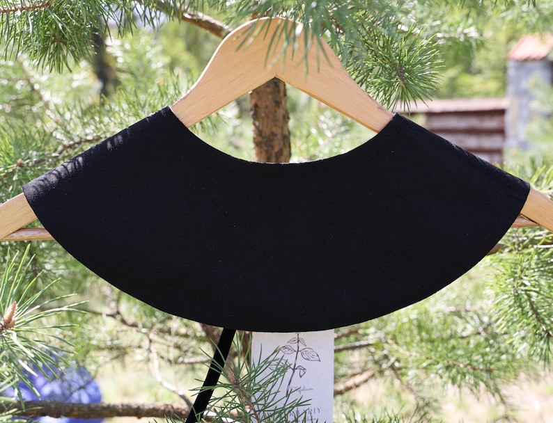 May include: A black, rounded collar, displayed on a wooden hanger. The collar has a smooth texture and a curved shape. The background features green foliage and a glimpse of a building. The collar is a fashion accessory.