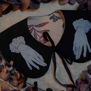 May include: A black collar with a floral patterned lining and a black ribbon tie. The collar features a silver-colored hand design on each side. The background includes dried leaves and a neutral-colored fabric.